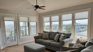 A living room with a grey sofa, ottoman, and armchairs, large windows with blinds, and a ceiling fan. - A Pirates Life - Fishing, Boat Slip, Pool, Gym!  (Manteo)