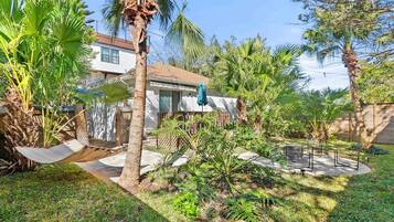 A residential backyard with a hammock, palm trees, a patio, and a house with a white exterior and brown roof.