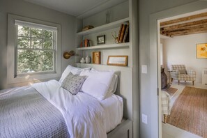 A bedroom with a large window, a bed with white linens, a bookshelf with books and decorative items, and a plaid armchair. - 18th Century Rosendale Cottage w/pool (Rosendale)