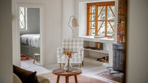 A cozy room with a plaid armchair, a small round table with a vase, a bookshelf, a fireplace, and a window with a view of trees. - 18th Century Cottage w/ heated pool 2 hrs from NYC (Rosendale)