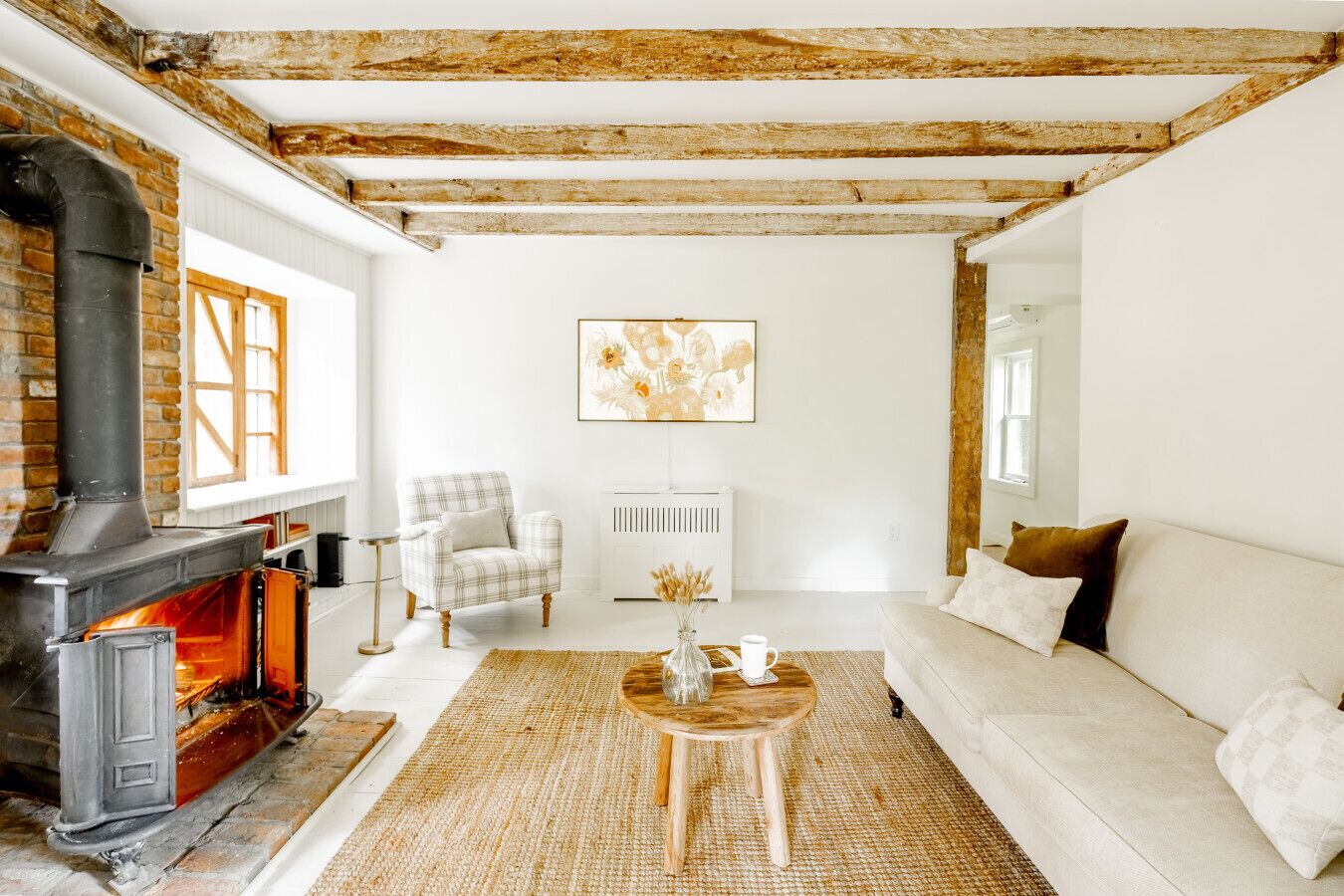 A cozy living room with exposed wooden beams, a fireplace, a white sofa, a plaid armchair, and a rug.