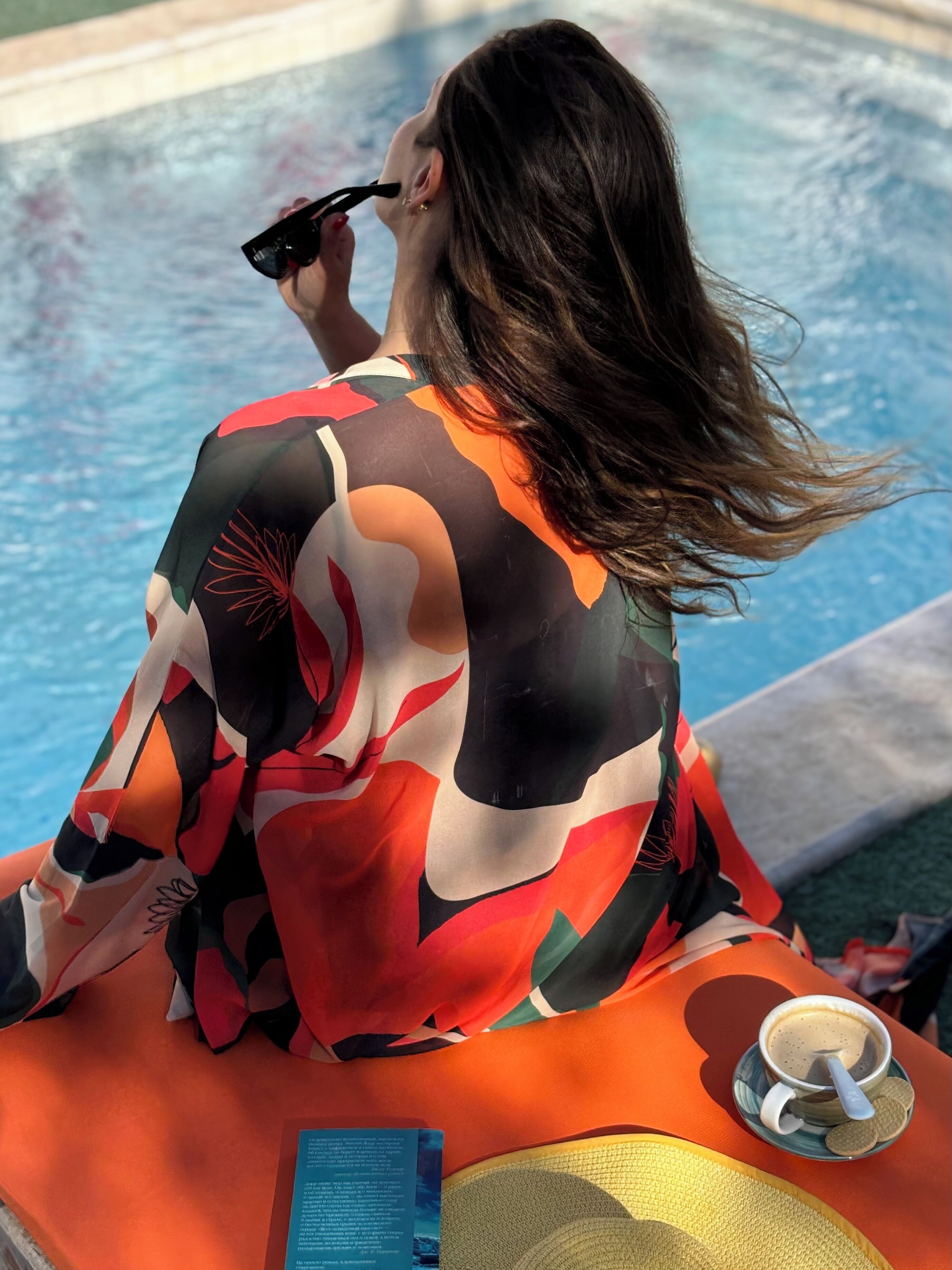 A person sitting by a pool with a colorful patterned garment, a cup of coffee, and a book on a yellow mat.