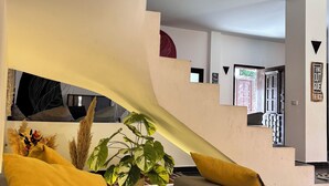 A modern living room with a grey sofa, yellow cushions, and a large plant. A staircase with a unique curved design leads to another level. - Charming American Villa   – 3BR with AC, WiFi & Pool in Dahab (Dahab)