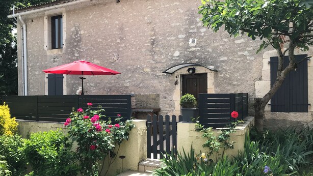A rustic stone house with a small porch, a red umbrella, and a gate.