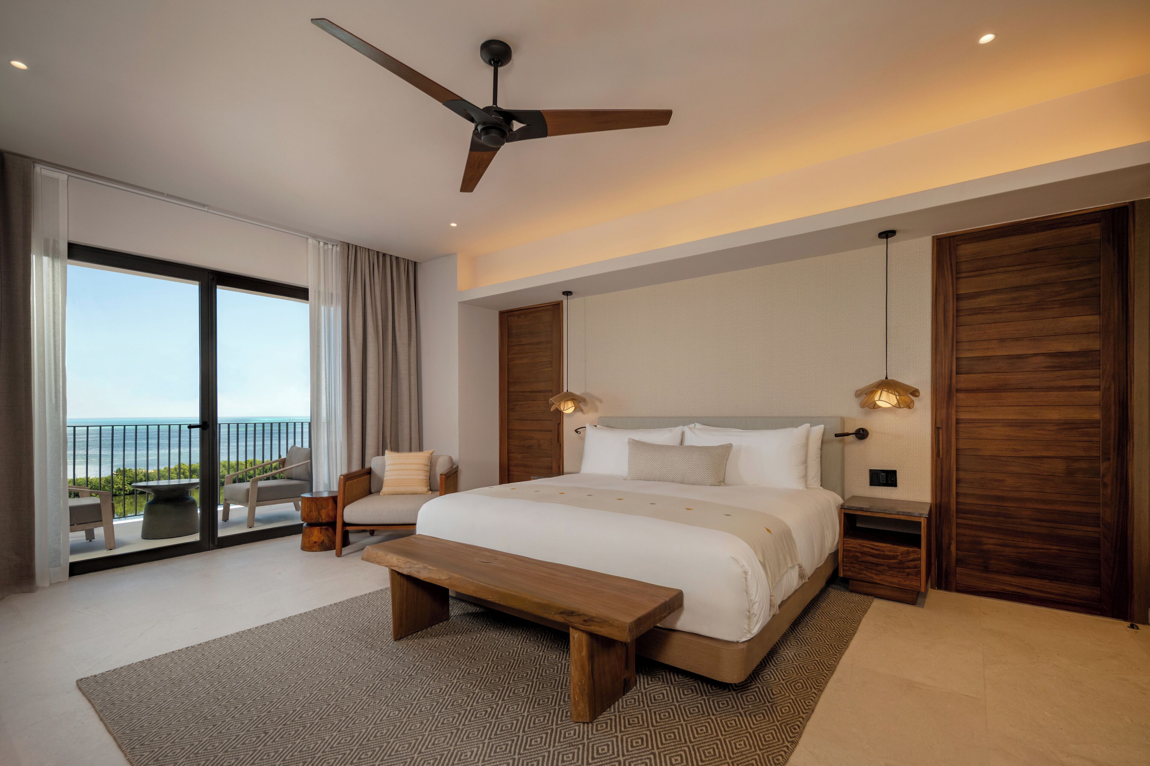 Un dormitorio moderno con una cama grande, un ventilador de techo, un banco y vistas al océano a través de amplios ventanales correderas.