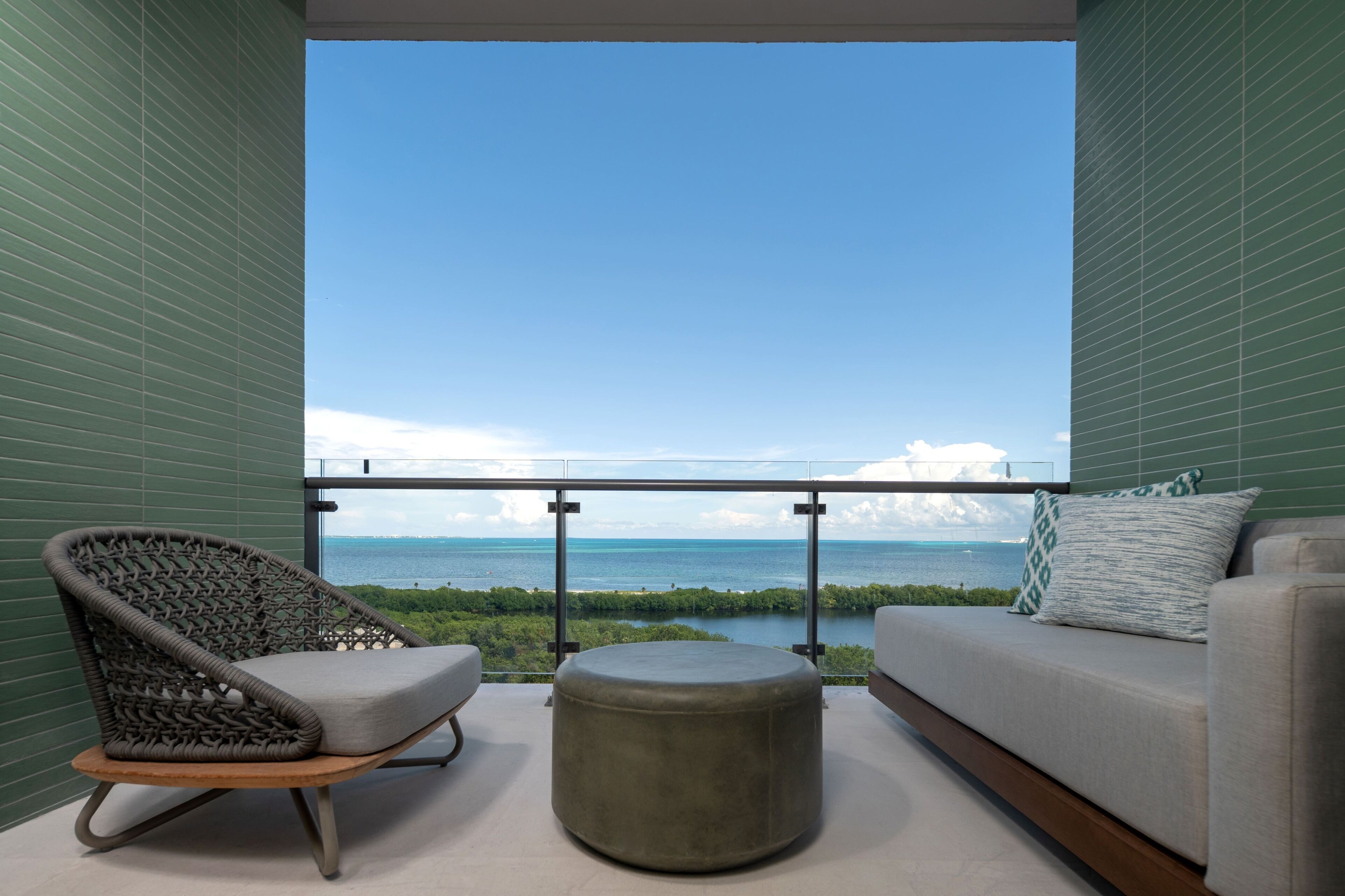 A balcony with a view of the ocean, a wicker chair, a round ottoman, and a sofa with cushions.