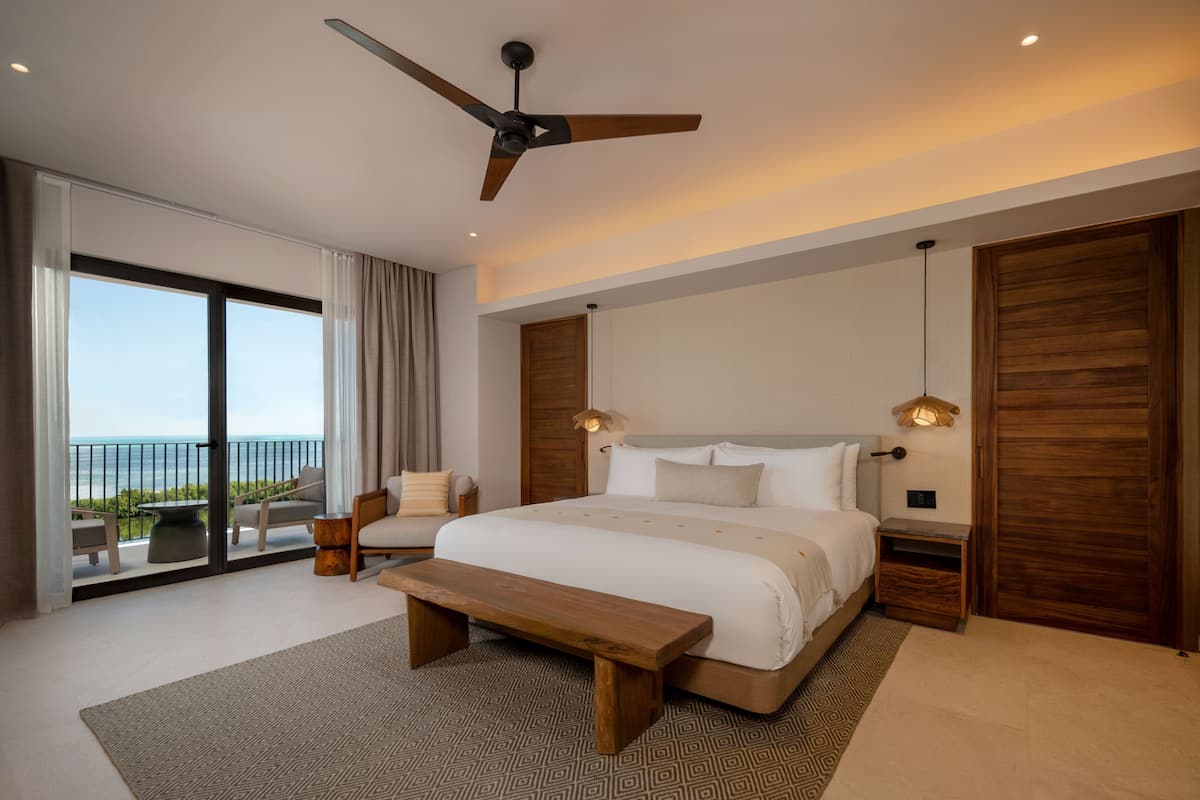 Een moderne slaapkamer met een groot bed, een plafondventilator, een houten bankje en uitzicht op zee door grote schuifdeuren.