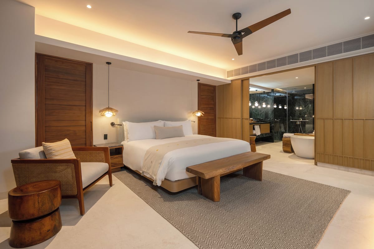 Een moderne slaapkamer met een groot bed, houten meubels, een plafondventilator en een badkamer die door een open deur zichtbaar is.
