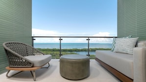 Een balkon met uitzicht op de oceaan, een rieten stoel, een rond voetenbankje en een bank met kussens.