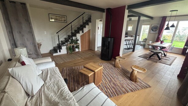 Amplia sala de estar con sofá, mesa de centro y chimenea. Se ve una escalera que conduce a otro nivel, y un comedor con mesa y sillas.
