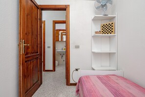A bedroom with a wooden door, a bed with a pink bedspread, a white radiator, and a shelf with a chessboard.