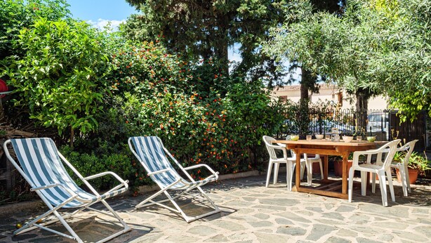 Un patio con suelo de piedra, dos sillones a rayas, una mesa de madera con sillas, y un fondo de follaje y árboles.