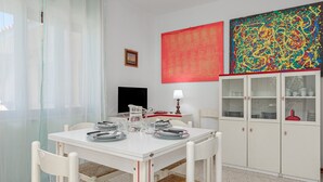 Un comedor con una mesa blanca, sillas de mimbre, un armario con puertas de cristal y obras de arte en las paredes.