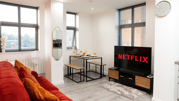 A modern living room with a red sofa, a dining table with benches, a black TV, and a clock on the wall. - Central Leicester Gem | Sleeps 5 | FREE PARKING (Leicester)