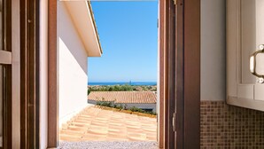 Vista a través de una puerta abierta que da a un balcón con suelo de baldosas y un cielo azul despejado.