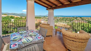 Un área de comedor al aire libre techada con una mesa para cuatro, sillas de mimbre y vistas al mar y al paisaje.