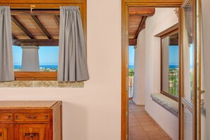A room with a wooden ceiling, a window with curtains, a wooden dresser, and a balcony with a view.