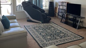 Una sala de estar con ventilador de techo, cinta de correr, televisor, estantería y un gran ventanal con cortinas.