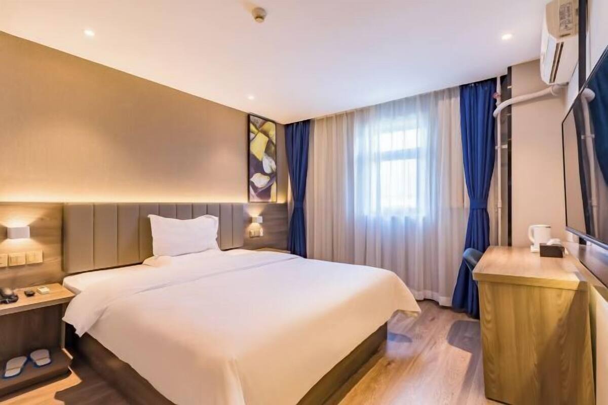 A hotel room with a bed, bedside table, lamp, and a small desk with a chair.