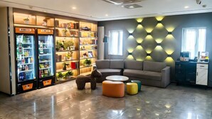 A modern hotel lobby with a sofa, a statue, a refrigerator with snacks, and a wall with geometric lighting.