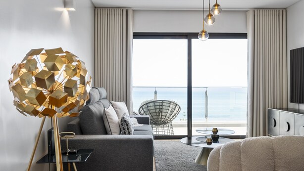 A modern living room with a sofa, a unique geometric floor lamp, a round chair, and a view of the sea through a large sliding glass door.