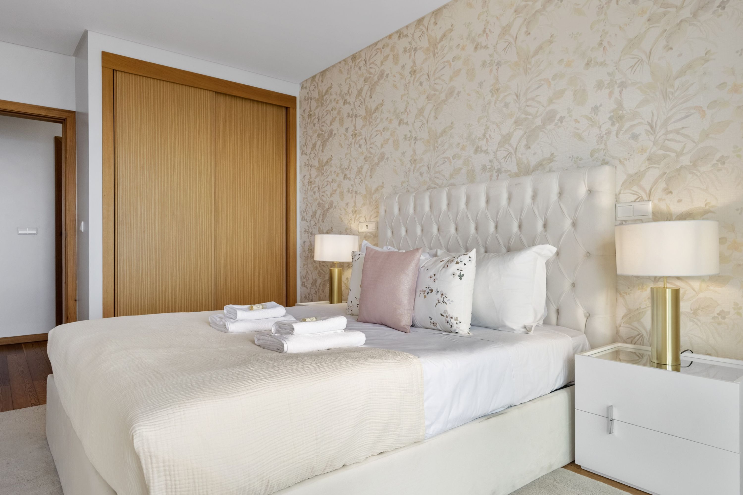Ein ordentlich bezogenes Bett mit weißer Decke und Kissen, ein Nachttisch mit Lampe sowie ein Holzschrank in einem Hotelzimmer.