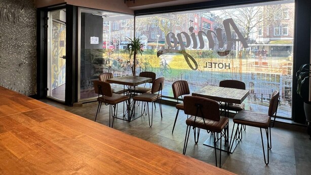 A modern restaurant with wooden tables and chairs, a large window with a view of the street, and a sign reading "HOPITAL HOTEL". - Amigo Hotel (Amsterdam)