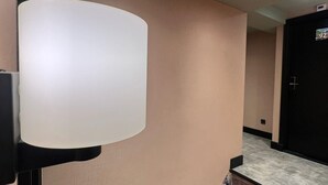 A hallway with a white wall, a security camera, a light fixture, and a door with a number sign. - Amigo Hotel (Amsterdam)