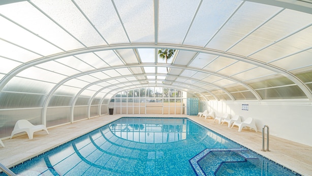 Ein Hallenbad mit Glasdach, klarem blauen Wasser und weißen Liegestühlen.