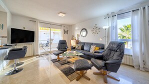 Ein modernes Wohnzimmer mit einer Couch, Sesseln, einem Couchtisch, einem Fernseher und einem großen Fenster.