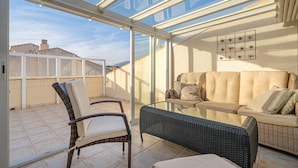 Eine sonnendurchflutete Terrasse mit Korbmöbeln, einer Glasüberdachung und Blick auf die Stadt.