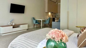 Moderni hotellihuone, jossa on sänky, seinälle asennettu televisio, ruokailualue tuoleineen ja pieni keittiönurkkaus.