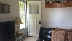 A living room with a black leather sofa, a television on a wooden stand, a lamp, and a wooden shelf above the sofa. - Cottage Retreat 75 Steps from the shores of spectacular Lake Huron, Sauble Beach (Sauble Beach)