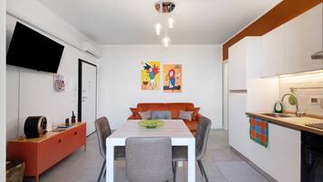 Een moderne keuken met witte kasten, een eettafel met stoelen, een flatscreen televisie aan de muur en twee kleurrijke kunstwerken.