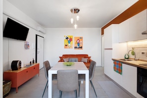 Une cuisine moderne avec des meubles blancs, une table à manger avec des chaises, un téléviseur à écran plat fixé au mur et deux œuvres d’art colorées accrochées au mur.