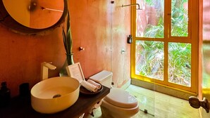 Une salle de bain avec un miroir rond, un lavabo, des toilettes et une douche. Il y a une fenêtre qui donne sur un paysage verdoyant.
