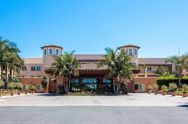 Un hôtel avec une entrée principale, des palmiers et un ciel d’un bleu éclatant.
