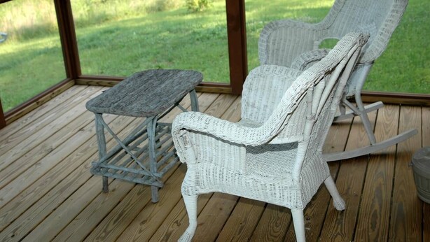 A wicker rocking chair and a small table on a wooden deck with a view of a grassy field.