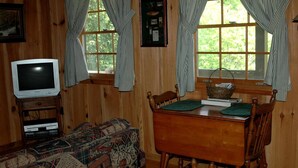 A cozy cabin room with a wooden interior, a television, a patterned sofa, a dining table with chairs, and large windows with curtains.