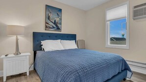 A bedroom with a blue bedspread, a painting of birds on the wall, a window with a view of a water tower, and a white nightstand with a lamp. - Cozy 1-bedroom condo in Lavallette, located 1 block from beach (Lavallette)