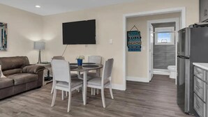 A modern living room with a grey sofa, a dining table with four chairs, a flat-screen TV mounted on the wall, and a kitchen area with a refrigerator and cabinets. - Cozy 1-bedroom condo in Lavallette, located 1 block from beach (Lavallette)