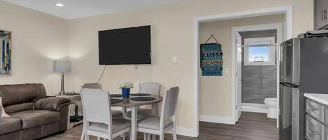 A modern living room with a grey sofa, a dining table with four chairs, a flat-screen TV mounted on the wall, and a kitchen area with a refrigerator and cabinets.