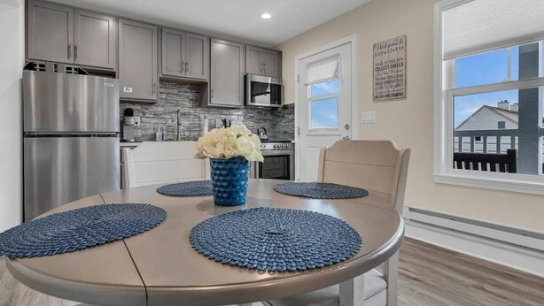 A modern kitchen with grey cabinets, stainless steel appliances, and a dining area with a round table and blue placemats. - Cozy 1-bedroom condo in Lavallette, located 1 block from beach (Lavallette)