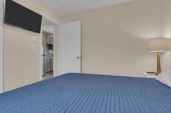 A bedroom with a blue bedspread, a white door, a kitchen in the background, and a bedside lamp. - Cozy 1-bedroom condo in Lavallette, located 1 block from beach (Lavallette)