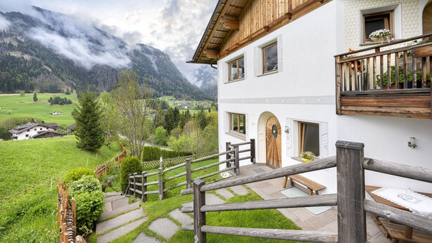 A white building with wooden balconies, a stone staircase, and a view of a green valley and mountains. - 1006A Apartments Ciancel Gino (Ortisei)
