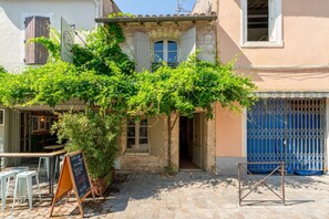 A cobblestone courtyard with outdoor seating, a signboard, and a building with a tiled roof and a blue gate.