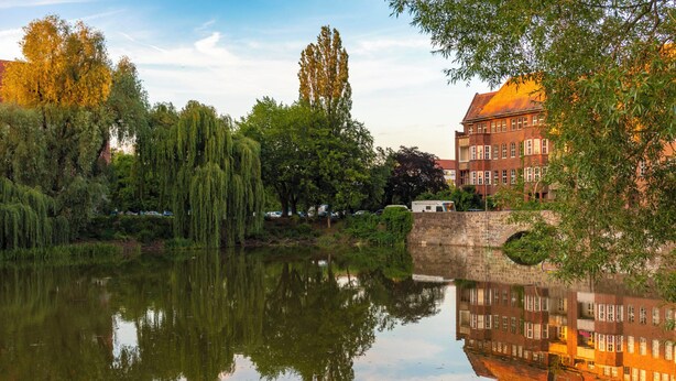 Ein ruhiger Fluss, der Bäume und ein Backsteinhaus mit rotem Dach spiegelt.