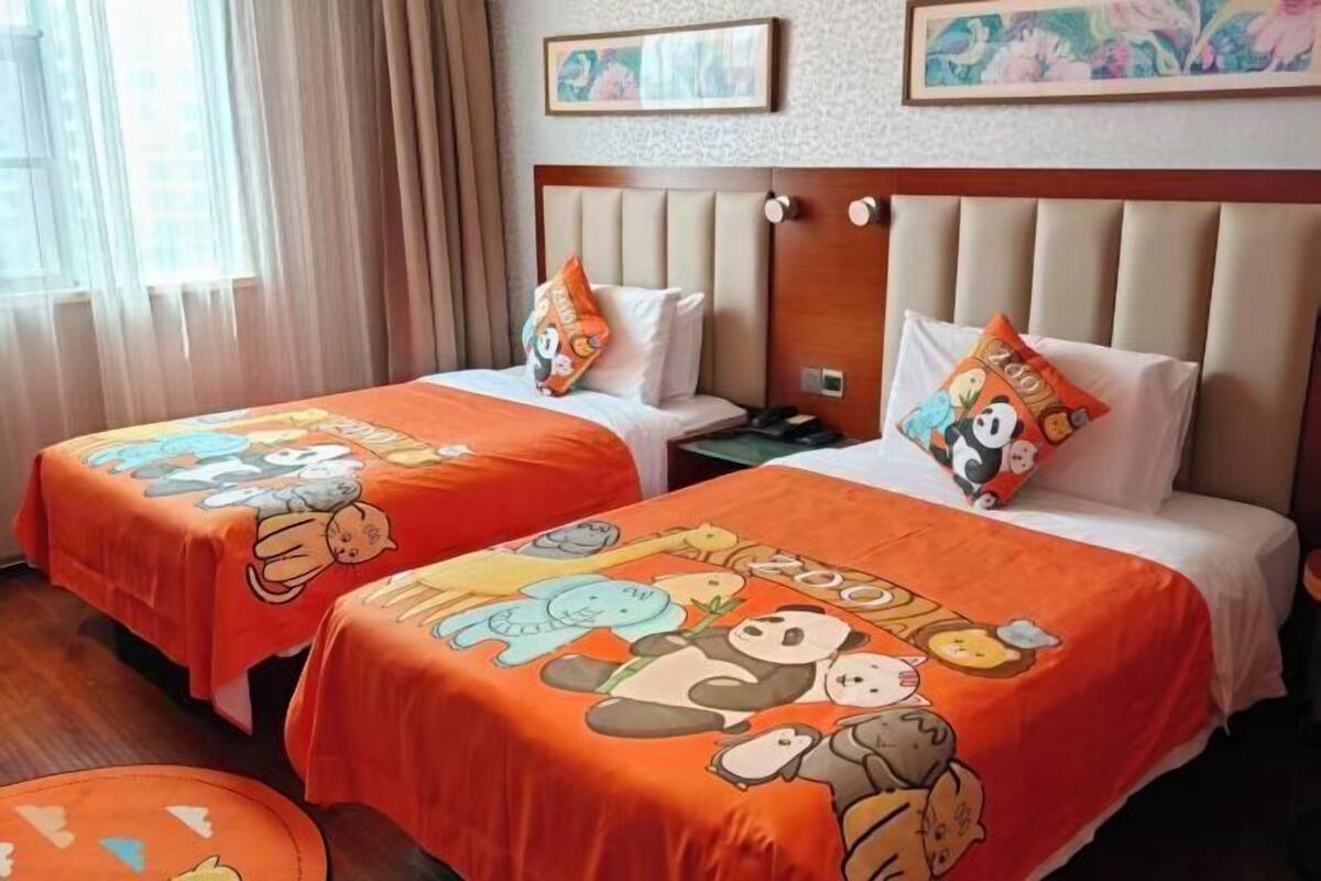 Quarto de hotel com duas camas, cada uma com colcha laranja decorada com personagens de desenho animado, uma cabeceira de madeira e uma mesa de cabeceira com abajur.