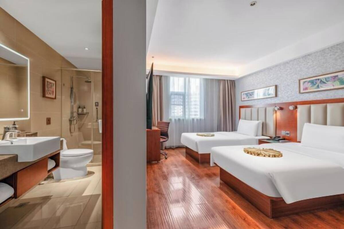 Quarto de hotel com piso de madeira, uma cama grande, mesa de cabeceira, escrivaninha, cadeira, banheiro com pia e vaso sanitário, e janela com cortinas.