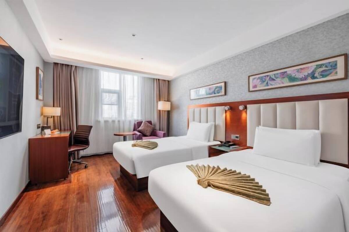 Quarto de hotel com duas camas, uma escrivaninha, uma cadeira, janela com cortinas e obras de arte nas paredes.
